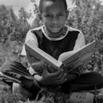 b&w photo young boy sitting in field cross-legged holding large open book, he is looking directly at the camera
